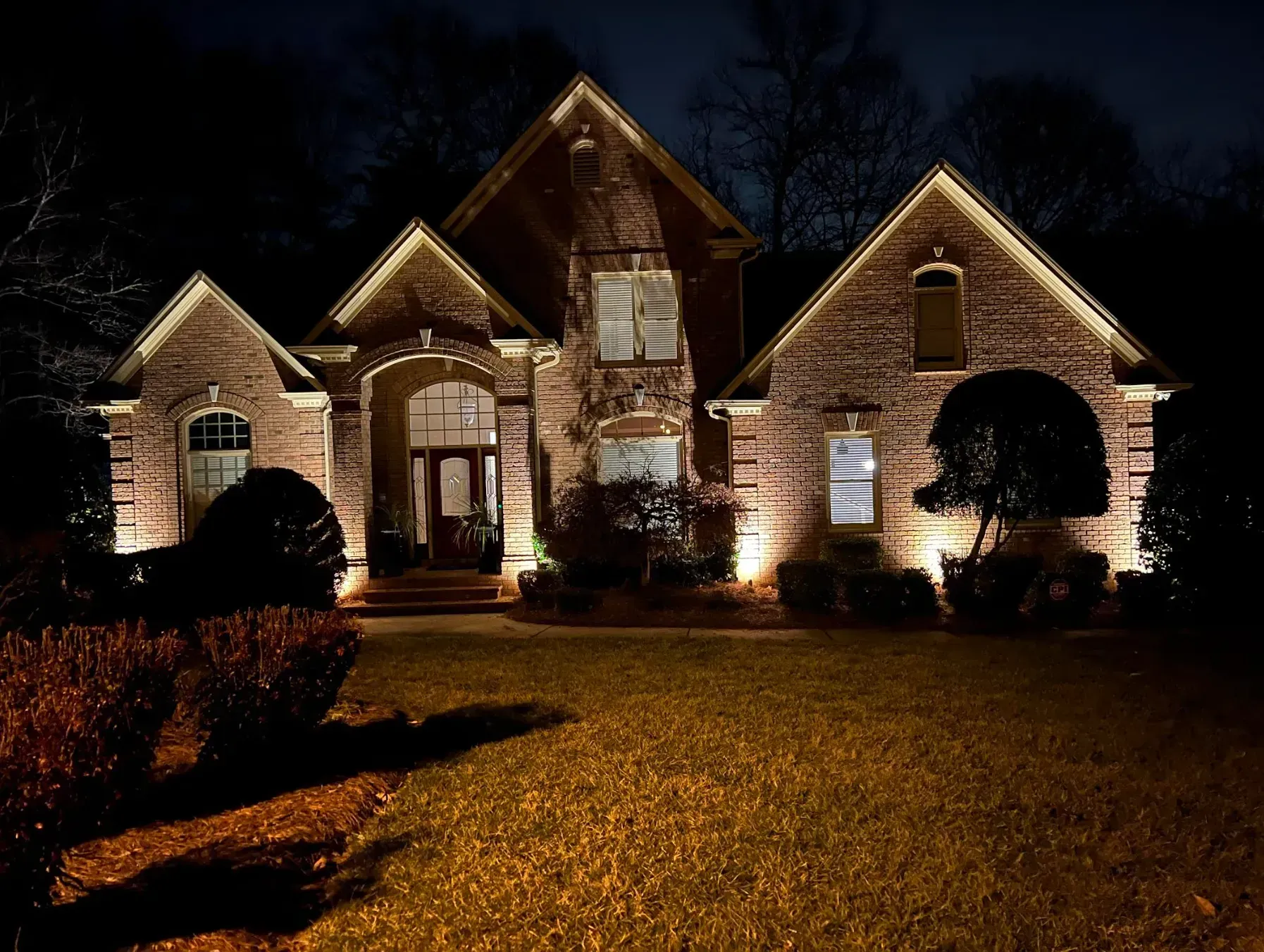 Brick house illuminated at night with spotlights on front facade and manicured lawn.